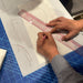 A close-up of hands using a KEARING brand, No 8002, Clear Plastic Straight Ruler, 50cm x 5cm, measuring with a pencil on a white drafting layout over a blue colour, Kai cutting mat, CB-143.